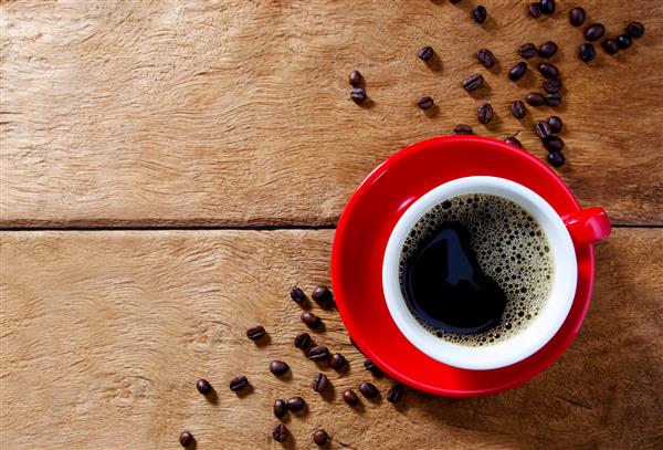 فنجان قهوه قرمز روی میز چوبی با دانه های قهوه پس زمینه با دانه های قهوه