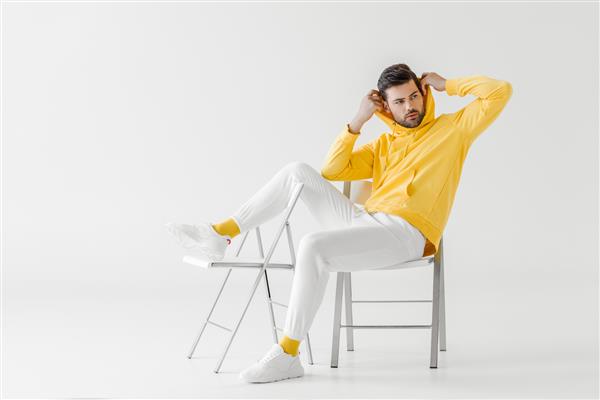 مرد جوان خوشتیپ با هودی زرد در حالی که روی صندلی های سفید نشسته است کاپوت می پوشد