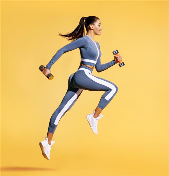 زن ورزشکار در حال پریدن با دمبل عکس زن فعال با لباس ورزشی در زمینه زرد حرکت پویا نمای کنار ورزش و سبک زندگی سالم