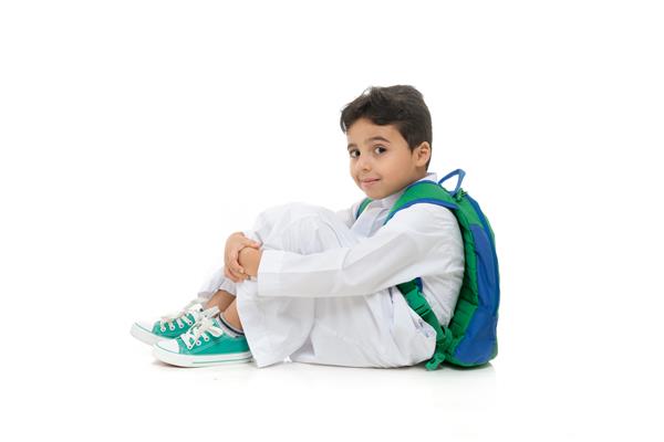 پسر مدرسه‌ای عرب که روی زمین نشسته با لبخندی بر صورت لباس سنتی عربستانی سفید کوله پشتی و کفش‌های کتانی به تن دارد و دستانش را روی پس‌زمینه جدا شده سفید بالا می‌برد
