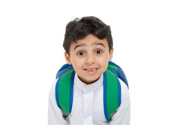 نمای نزدیک پسر مدرسه‌ای عرب روی صورت با لبخند و دندان شکسته با لباس سنتی عربستانی سفید کوله پشتی و کفش‌های کتانی دستانش را روی پس‌زمینه جدا شده سفید بالا می‌برد