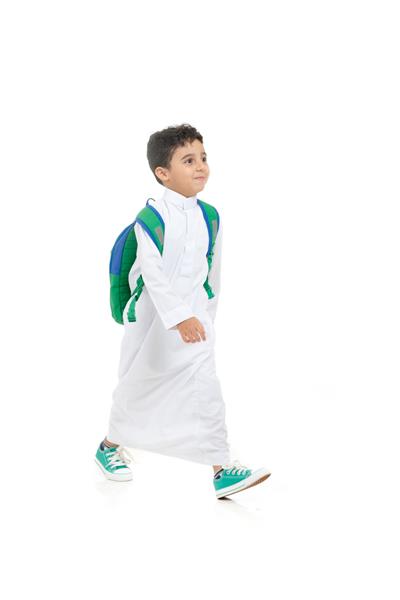 پسر مدرسه‌ای عرب لبخند می‌زند و راه می‌رود با لباس سنتی عربستانی سفید کوله پشتی و کفش‌های کتانی دست‌هایش را روی پس‌زمینه جدا شده سفید بالا می‌برد