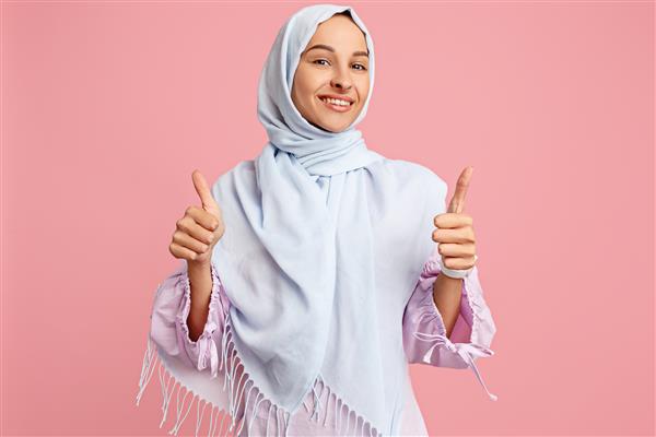 زن عرب شاد با حجاب با علامت ok پرتره دختر خندان که در پس زمینه صورتی استودیو ژست گرفته است زن جوان احساساتی احساسات انسانی مفهوم حالت چهره نمای جلویی