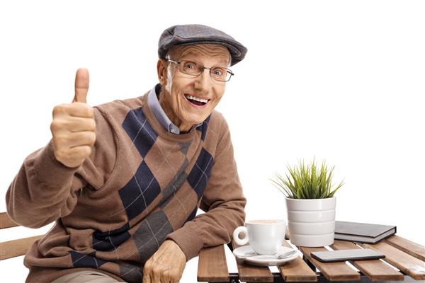 سالمندی که پشت میز قهوه نشسته است با ژست انگشت شست رو به بالا در پس زمینه سفید جدا شده است