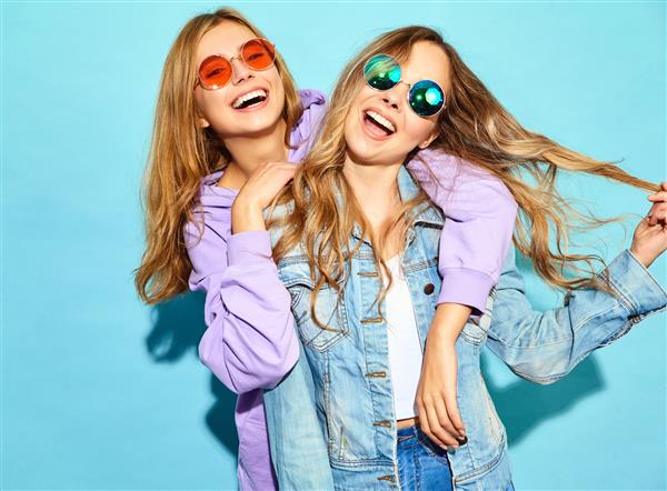 دو دختر جوان بور و خندان با لباس های تابستانی مد روز زنان بی خیال سکسی که در نزدیکی دیوار آبی با عینک آفتابی ژست گرفته اند مدل های مثبت در حال تفریح