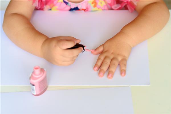 دختر بچه شایان ستایشی که سرگرم بازی در خانه با لاک صورتی رنگارنگ در حال انجام مانیکور و نقاشی ناخن است کودک نوپا ناز با لباس صورتی زیبا روی میز سفید چوبی نشسته و مانیکور درست می کند
