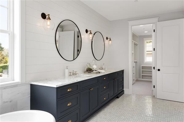 حمام زیبای سفید و روشن در خانه لوکس جدید با کابینت های روشویی بزرگ و آبی تیره دارای وان حمام و دو سینک و آینه های مدور کمد دیواری را نشان می دهد