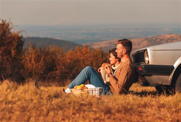 زوج جوان زیبا که از زمان پیک نیک در غروب آفتاب لذت می برند آنها چای می نوشند و در یک چمنزار می نشینند و به یک ماشین قدیمی تکیه داده اند