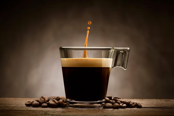 قهوه سیاه در فنجان شیشه ای با دانه های قهوه و قطره پرش روی میز چوبی