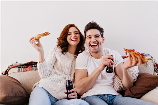 زن و مرد خندان در حال خوردن پیتزا هنگام تماشای فیلم عکس داخل خانه از زوج خوشبختی که از آخر هفته لذت می برند