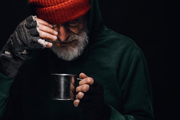 پیرمرد بی خانمان با ریش خاکستری پوشانده شده با لباس های نازک سبز در حالی که یک فنجان چای داغ در دست دارد تا خود را در یک شب سرد گرم کند