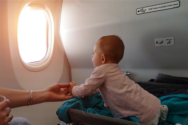 نوزاد خوشحال در حوض مخصوص در هواپیما در شکم خود می خوابد اولین پرواز کودک او تحت تاثیر قرار گرفته و به پنجره هواپیما نگاه می کند