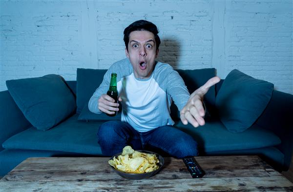 مرد جوان در حال نوشیدن آبجو در خانه در حال تماشای مسابقه فوتبال فوتبال یا بازی ورزشی در تلویزیون در شب فریاد زدن و جشن گرفتن گل یا پیروزی در جشن ورزش و مفهوم طرفداران