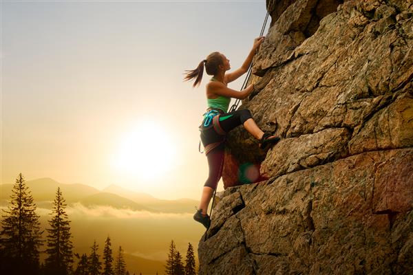 زن زیبا در حال بالا رفتن از صخره بلند در غروب مه آلود در کوهستان مفهومی ماجراجویی و ورزش شدید
