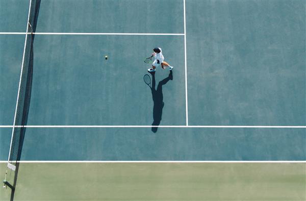 نمای هوایی از تنیس باز مرد جوان که در زمین سخت برای برداشتن توپ راه می رود تنیس باز حرفه ای در زمین باشگاه