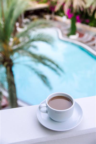 یک فنجان قهوه با شیر در یک کاسه سفید در بالکن هتل مشرف به آب آبی استخر در میان درختان نخل زمینه