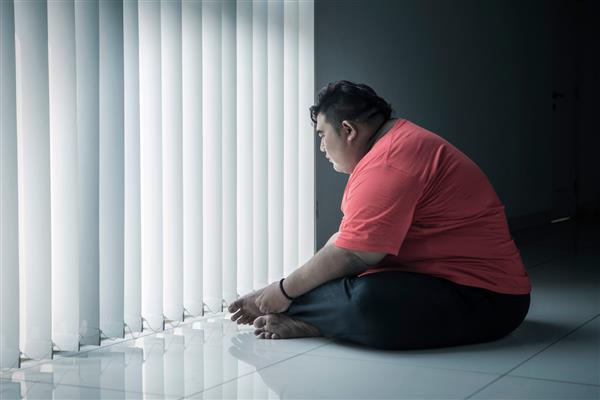 تصویر مرد چاق جوان در حالی که نزدیک پنجره نشسته متفکر به نظر می رسد