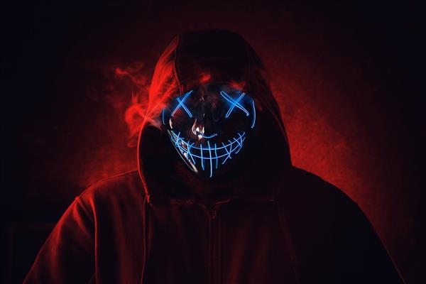 مردی با ماسک نئونی نورپردازی خشمگین و ترسناک در کاپوت روی پس‌زمینه قرمز تیره مفهوم هالووین و ترسناک