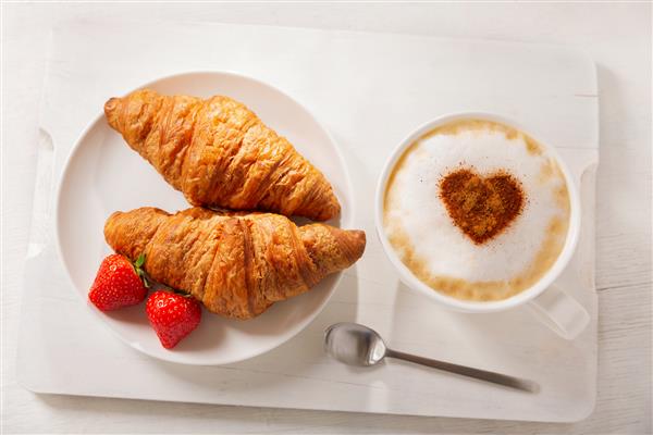 روز ولنتاین فنجان قهوه کاپوچینو با قلب کشیده و کروسان های تازه روی تخته چوبی نمای بالا