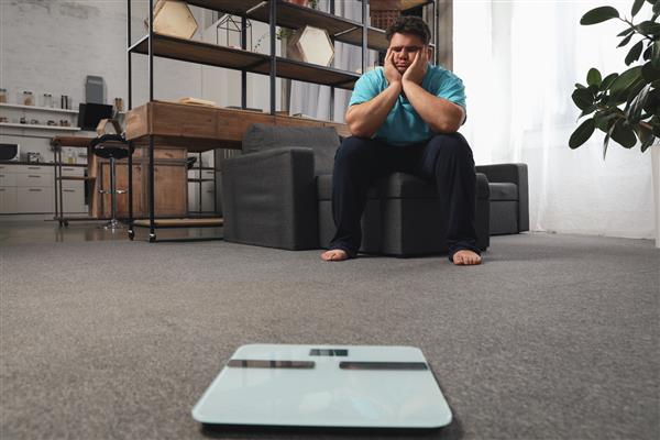 ترازو و مرد افسرده اضافه وزن روی زمین در خانه