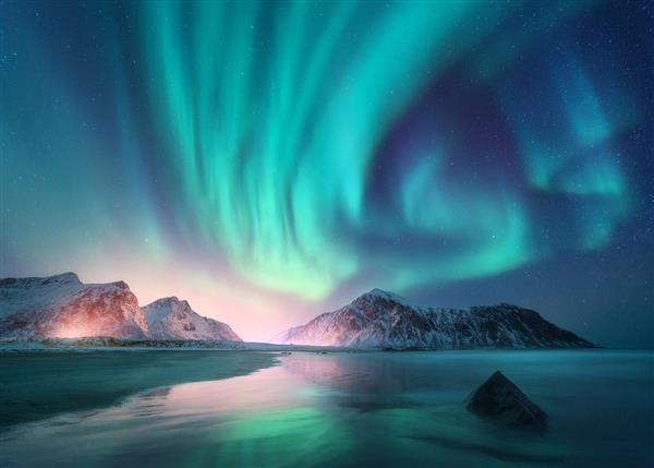 شفق قطبی بر فراز دریا کوه های برفی و نور شهر در شب شفق شمالی در جزایر لوفوتن نروژ آسمان پرستاره با نورهای قطبی منظره زمستانی با شفق قطبی انعکاس ساحل شنی