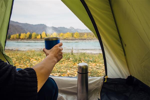 لیوان کمپر با چای داغ داخل چادر سبز در پس زمینه منظره کوهستانی زیبا نمای از داخل