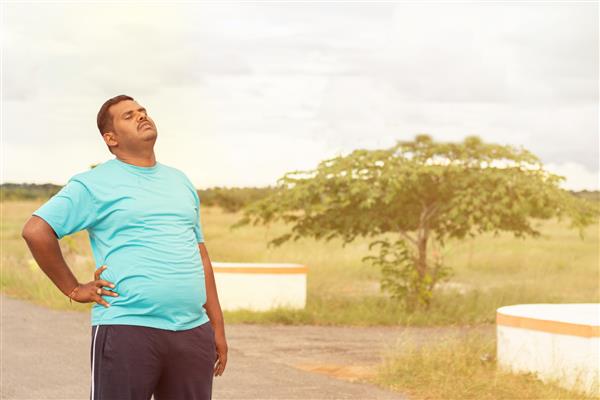 مرد جوان چاق خسته که پشت خود را در حین دویدن نگه می دارد - مفهوم تناسب اندام مرد چاق و سبک زندگی ناسالم