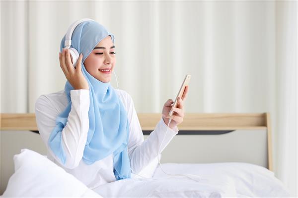 پرتره زن مسلمان آسیایی زیبا با لباس خواب در حال تماشای داستان آنلاین با تلفن همراه لایت روی تخت و متصل به اینترنت بی سیم دختر جوان ناز با حجاب گوش دادن به موسیقی از گوشی هوشمند