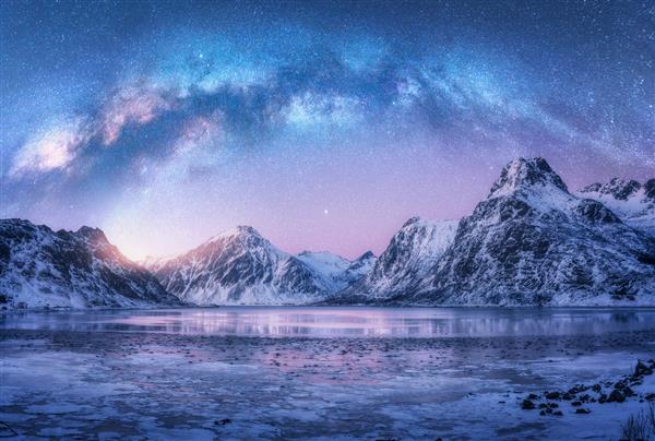 راه شیری در بالای ساحل دریای یخ زده و کوه های پوشیده از برف در زمستان در شب در جزایر لوفوتن نروژ منظره قطب شمال با آسمان پر ستاره آبی آب یخ سنگ های برفی راه شیری فضا و کهکشان