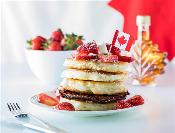 نمای نزدیک از یک پشته پنکیک و توت فرنگی برای روز کانادا با یک بطری شربت برگ افرا مفهوم روز کانادا