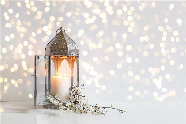 گل‌های سفید شکوفه‌های درخت آلو و فانوس نقره‌ای درخشان تزئینی مراکشی روی پس‌زمینه میز با چراغ‌های بوکه طلایی شام افطار کارت تبریک رمضان کریم دعوتنامه