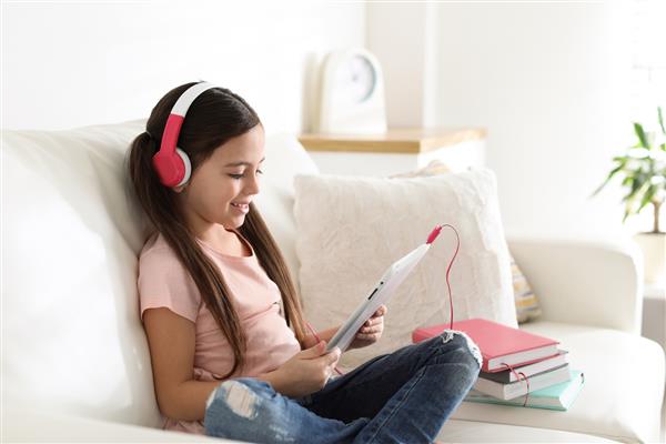 دختر بچه ناز با هدفون و تبلت در حال گوش دادن به کتاب صوتی در خانه