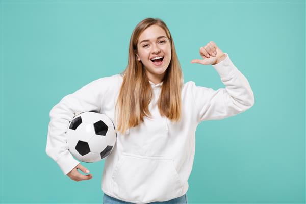 دختر جوان بامزه هوادار فوتبال با هودی سفید جدا شده روی زمینه آبی فیروزه ای مفهوم سبک زندگی تفریحی ورزشی تیم مورد علاقه خود را با توپ فوتبال که انگشت شست را به سمت خود گرفته است تشویق کنید