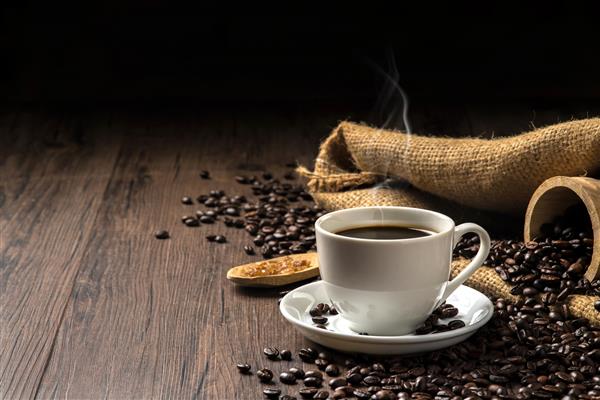 قهوه داغ در یک فنجان قهوه سفید و تعداد زیادی دانه قهوه در اطراف و شکر روی میز چوبی در فضایی گرم و روشن در زمینه تیره با فضای کپی