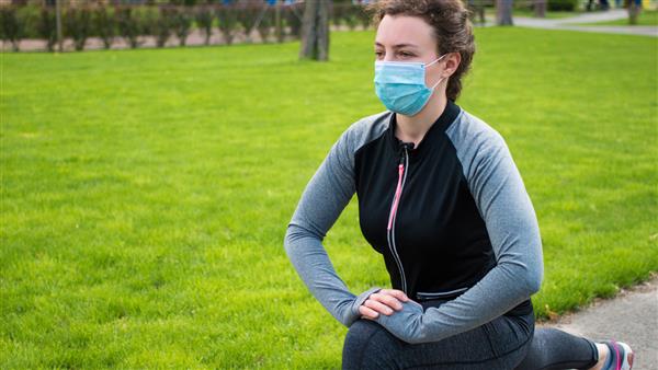 ورزش در دوران قرنطینه ویروس کرونا کووید-19 زن جوان ورزشکار با ماسک محافظ پزشکی که پاهایش را بعد از دویدن در فضای باز دراز می کند دختر تناسب اندام در حال انجام ورزش لانگز تمرین تمرین