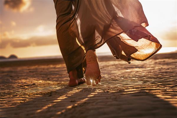 نمای نزدیک از پاهای زن در حال راه رفتن در ساحل هنگام غروب آفتاب