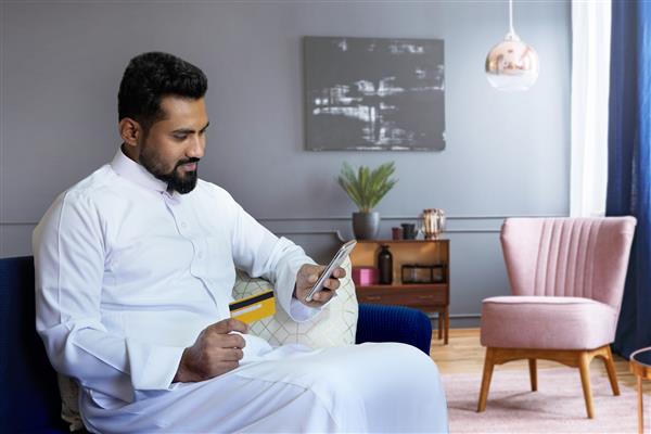 مرد سعودی در حال پرداخت آنلاین در خانه و نشستن روی مبل در اتاق نشیمن