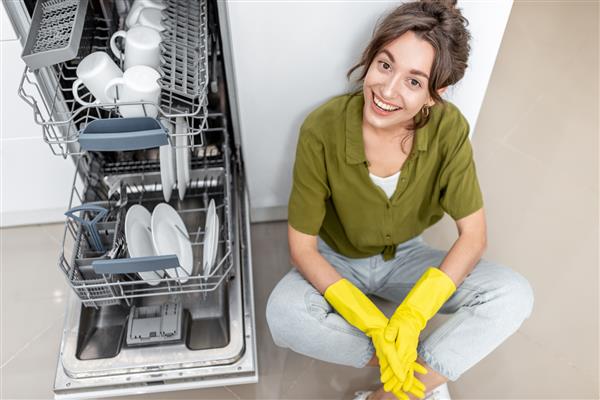 پرتره یک زن خانه دار خوشحال که در نزدیکی ماشین ظرفشویی با ظروف تمیز روی آشپزخانه خانه نشسته است کار آسان خانه با مفهوم لوازم آشپزخانه