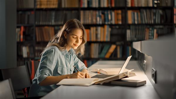 کتابخانه دانشگاه دختر زیبای قفقازی باهوش از لپ‌تاپ استفاده می‌کند یادداشت‌هایی را برای کاغذ انشا مطالعه برای تکالیف کلاس می‌نویسد یادگیری دانشجویان متمرکز مطالعه برای امتحانات کالج پرتره نمای جانبی