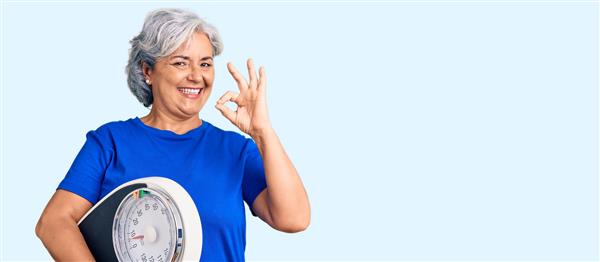 زن سالخورده با موهای خاکستری که وزنه را نگه می‌دارد برای متعادل کردن کاهش وزن علامت ok را با انگشتان انجام می‌دهد لبخند دوستانه با اشاره نماد عالی