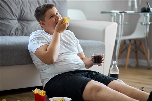 پسر نوجوان چاق دارای اضافه وزن تغذیه بدی دارد غذای ناسالم بخورد روی زمین بنشینید و غذای ناسالم بخورید و تلویزیون تماشا کنید