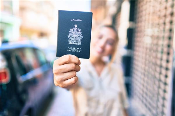 زن جوان و زیبای بلوند قفقازی که در یک روز آفتابی در فضای باز لبخند می زند و پاسپورت کانادا را نشان می دهد