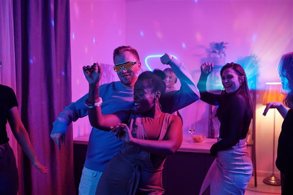 گروهی از دوستان جوان شاد بین فرهنگی با لباس‌های غیررسمی هوشمند در حال رقصیدن هیجان‌زده در مهمانی خانگی در اتاق نشیمن با نورپردازی صورتی