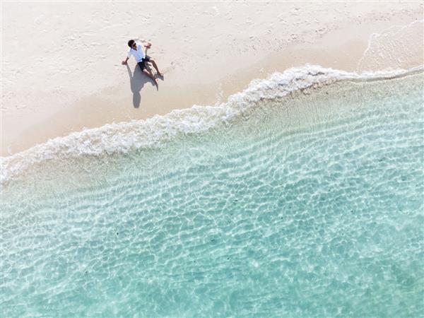 مرد جوانی نشسته و در حال استراحت در ساحل زیبای استوایی ماسه سفید با فوم موج و دریای شفاف تعطیلات تابستانی و پس‌زمینه سفر نمای بالا از پهپاد