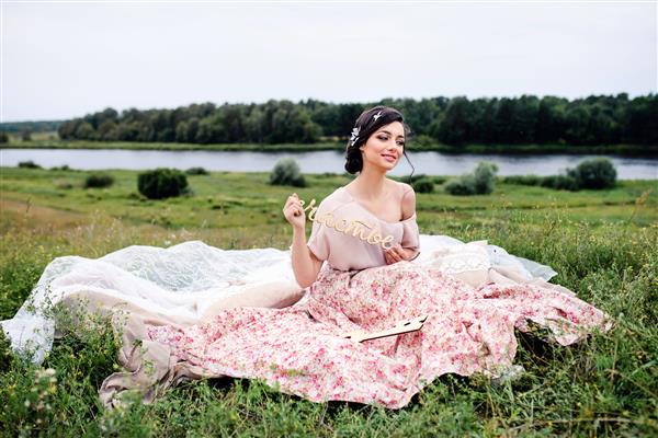زن زیبای سبزه با لباسی که روی چمن نزدیک رودخانه نشسته است کتیبه چوبی به معنای خوشحالی است