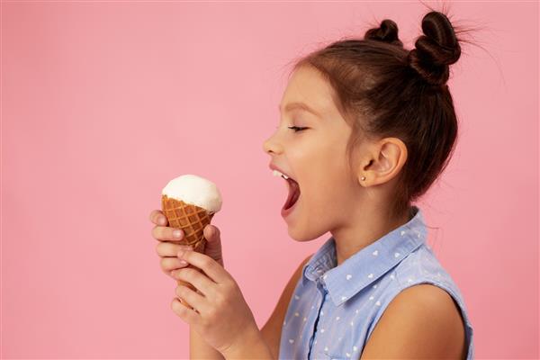 دختر کوچک خندان بامزه در حال خوردن بستنی وانیلی در مخروط وافل در زمینه صورتی