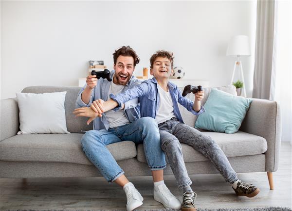 پدر و پسری شاد با جوی استیک در حال بازی ویدیویی در خانه پسری که با دست حواس پدر را پرت می کند