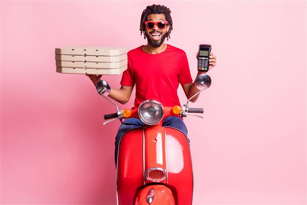 پرتره پسر خوش تیپ و خوش تیپ سوار بر موتور سیکلت در حال تحویل پیتزا در دست ترمینال پرداخت بی سیم جدا شده روی پس زمینه صورتی رنگ
