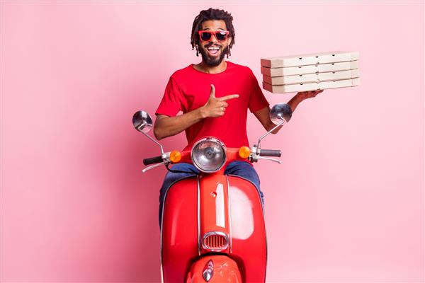 پرتره پسر خوش تیپ و متحیر سوار بر موتور سیکلت در حال تحویل تبلیغ پیتزا که روی پس زمینه رنگی پاستلی صورتی جدا شده است
