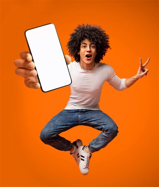 پرتره پسر نوجوان آفریقایی آمریکایی هیجان زده در حال پریدن با گوشی هوشمند نمایش صفحه خالی در پس زمینه نارنجی فضای کپی برای تبلیغات موبایل شما تصویر ماکت